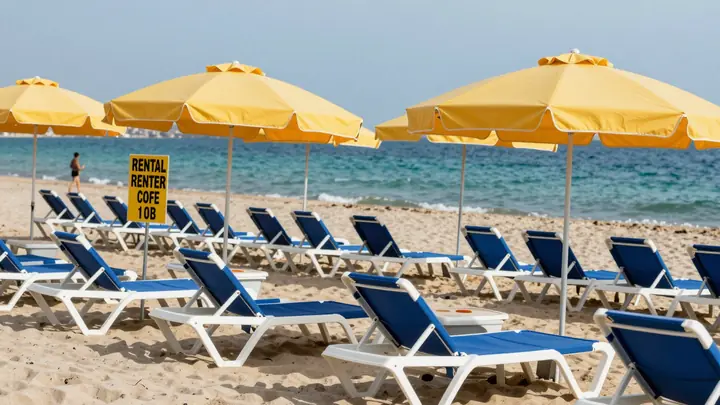 Rows of blue and white beach loungers with yellow umbrellas on a sunny city beach, turquoise sea in background, rental s